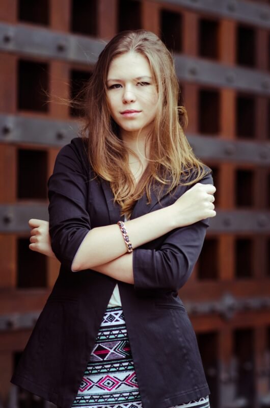 Portrait of a young woman with light brown hair wearing a black blazer and patterned skirt posing with arms crossed