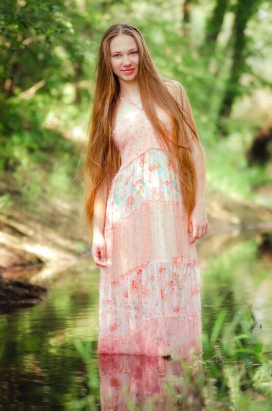 Smiling young woman with extremely long hair wearing a pink floral maxi dress standing in a shallow forest river