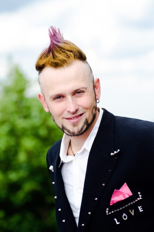 Smiling man with a creative punk mohawk hairstyle, wearing a black blazer with a pink pocket square and the word LOVE.