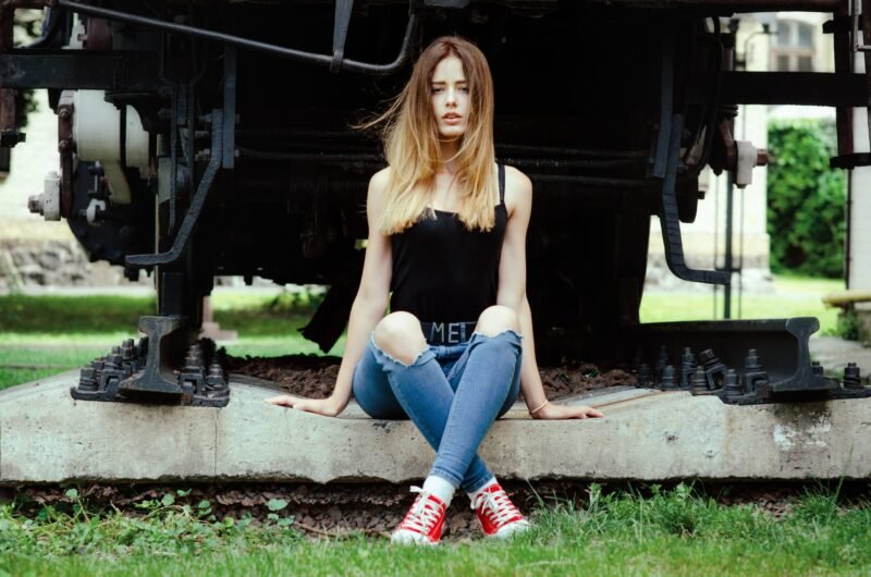 Full length portrait of a young woman in a black top and ripped jeans sitting on a train track platform