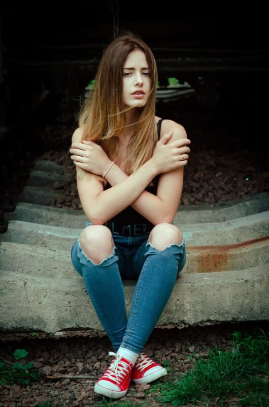 Young blonde woman sitting with arms crossed wearing a black tank top, ripped jeans, and red sneakers