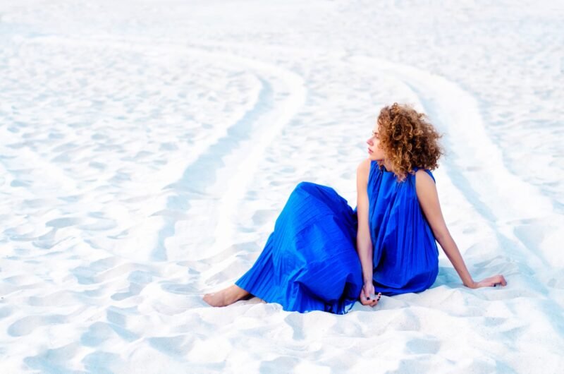 Woman in a vibrant blue dress sitting on a white sand beach looking away