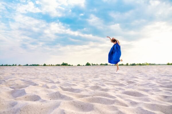 Distant view of a woman in a flowing blue dress jumping joyfully on a white sand desert under a cloudy sky