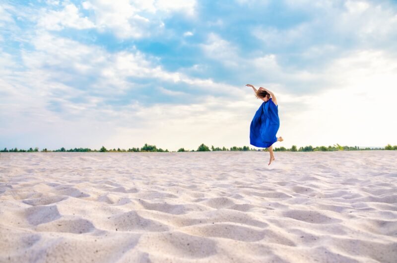 Distant view of a woman in a flowing blue dress jumping joyfully on a white sand desert under a cloudy sky