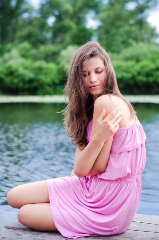 Full length portrait of a young woman in a pink dress sitting on a wooden pier by a lake surrounded by green trees.