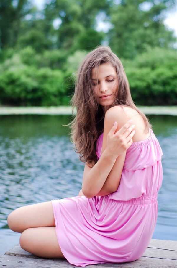 Full length portrait of a young woman in a pink dress sitting on a wooden pier by a lake surrounded by green trees.