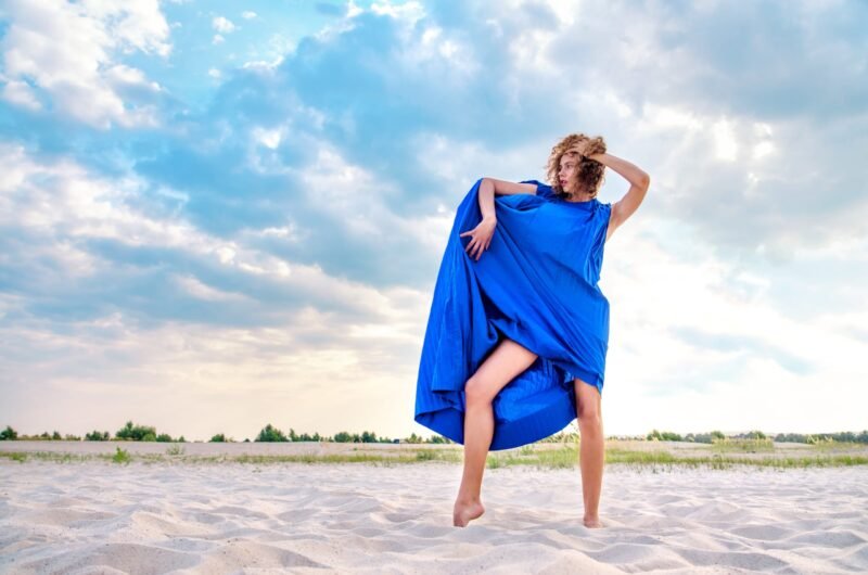 Woman in a bright blue dress posing dynamically on a white sand beach under a blue sky