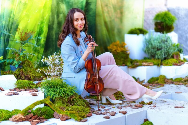 A woman in a blue blazer and pink trousers sitting down, holding a violin in a decorative green garden setup.
