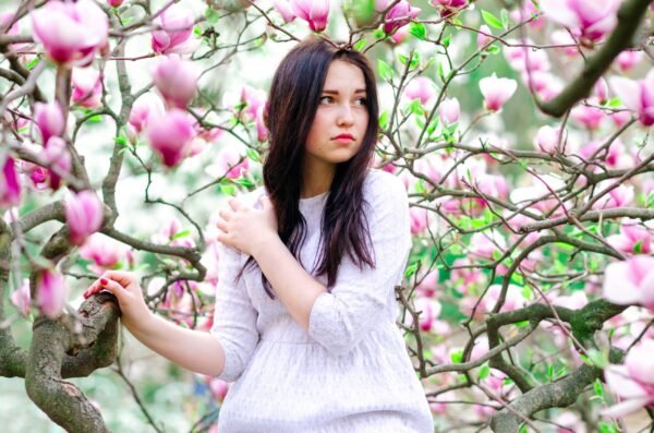 A young woman with long dark hair posing thoughtfully amidst blooming pink magnolia trees in spring.