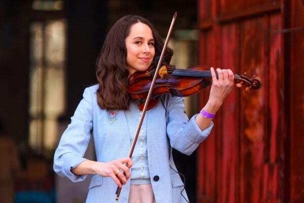 A smiling female violinist in a light blue blazer playing the violin outdoors against a rustic red wooden wall.