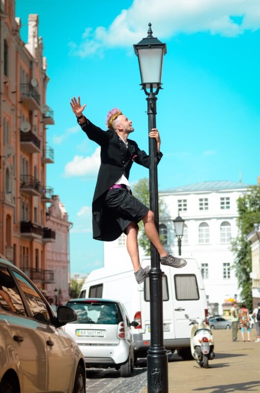 A man with pink-tipped hair in a black coat climbing a classic street lamp post in a city square under a bright blue sky.