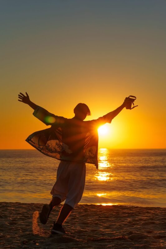 Silhouette of a person with outstretched arms running joyfully on a sandy beach during a vibrant golden sunset.