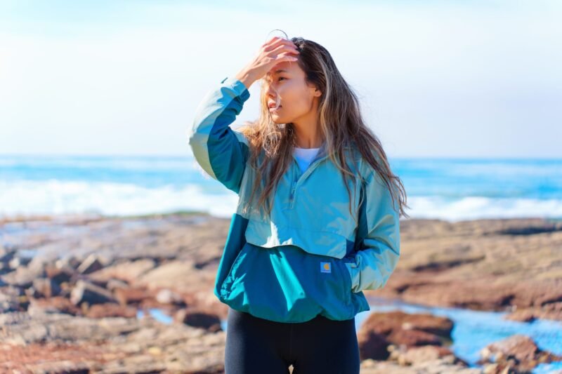 A young woman in a turquoise windbreaker adjusting her hair while standing on a rocky coastline with the ocean in the background.