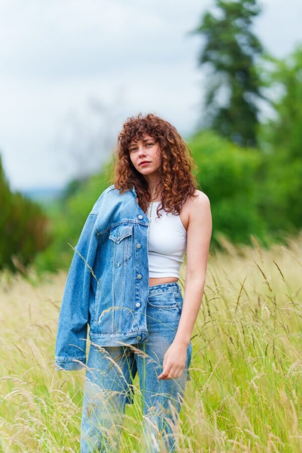 A young woman with curly brown hair standing in a grassy field with a denim jacket draped over her shoulder.