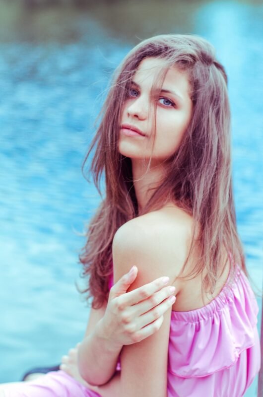 Young woman with long brown hair wearing a pink dress, looking at the camera while posing outdoors by the blue water.