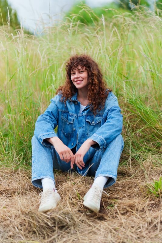 A smiling young woman with curly hair wearing a denim jacket and jeans, sitting happily in a field of tall grass.