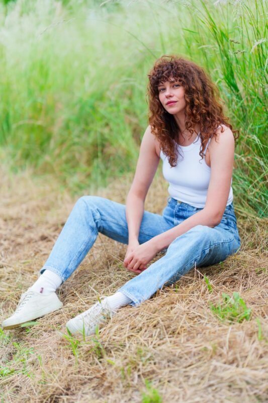A young woman with curly brown hair and bangs sitting on dry grass in a field, wearing a white tank top, blue jeans, and white sneakers.