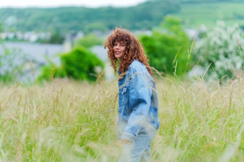 A smiling woman with curly brown hair looking back over her shoulder while walking through a field of tall grass in a denim jacket.