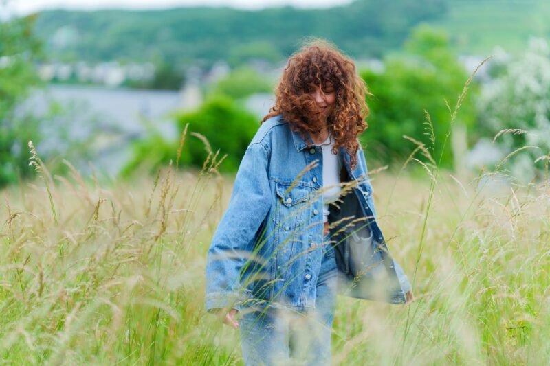 A young woman with curly hair walking through a tall green grass field while wearing an oversized blue denim jacket.