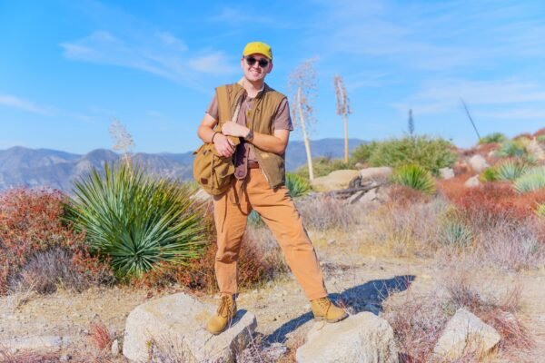 A smiling man wearing a yellow cap, sunglasses, brown vest, and khaki pants stands on rocks in a desert mountainous landscape.