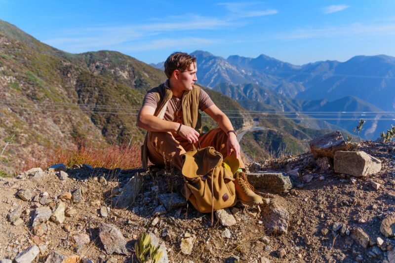 A young man sitting on a rocky hillside next to a canvas backpack, looking out over a vast mountain range under a clear blue sky.