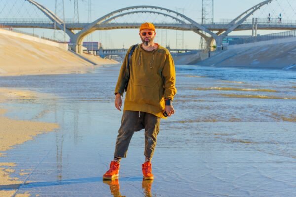A man wearing an orange cap, sunglasses, olive green sweatshirt, and bright red sneakers stands in a shallow concrete water canal with a large bridge in the background.