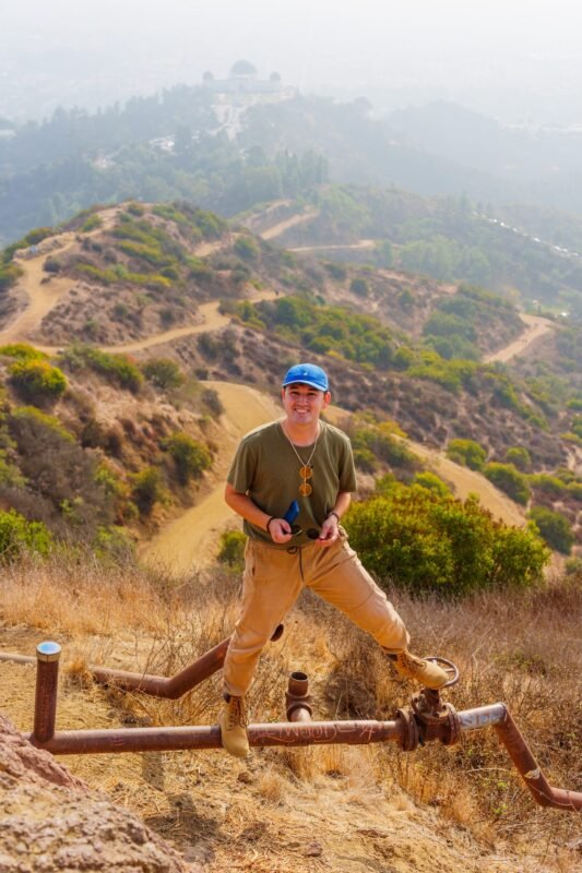 A young man wearing a blue baseball cap and olive green t-shirt stands on a metal pipe on a hillside overlooking foggy mountains.