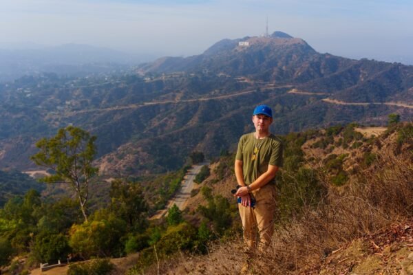 A man wearing a blue cap stands on a hillside trail with the Hollywood hills and a communication tower visible in the hazy distance.