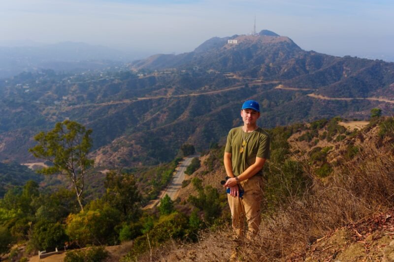 A man wearing a blue cap stands on a hillside trail with the Hollywood hills and a communication tower visible in the hazy distance.