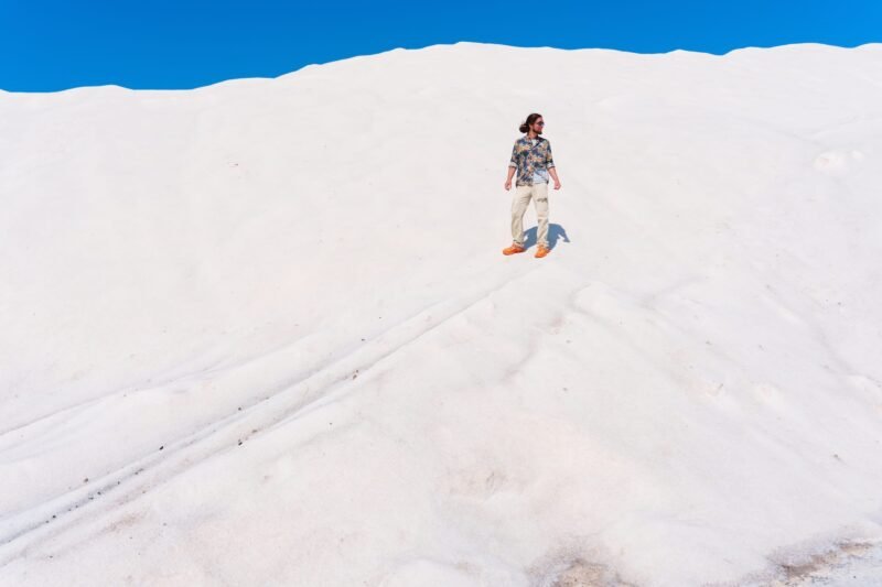 Distant view of a man with long hair standing on a massive white sand dune under a bright blue sky.