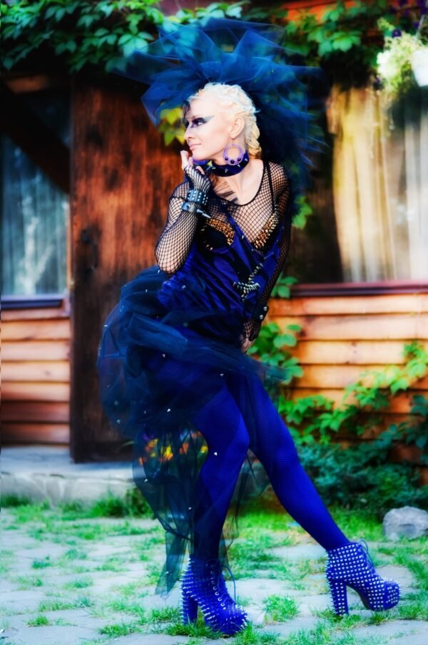 Woman in an edgy punk gothic style outfit with blue tights, studded boots, and dramatic black tulle headpiece posing outdoors.