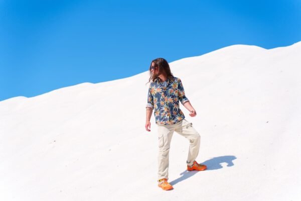 A man with long hair wearing a floral patterned shirt and bright orange shoes walks up a bright white sand dune against a clear blue sky.