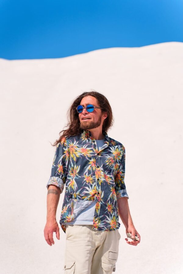 A man with long hair and a beard wearing blue sunglasses and a tropical floral shirt, standing against a white sand dune and clear blue sky.