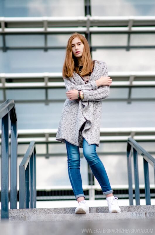 A young woman with red hair posing on concrete stairs, dressed in a grey textured cardigan, blue denim jeans, and white shoes.