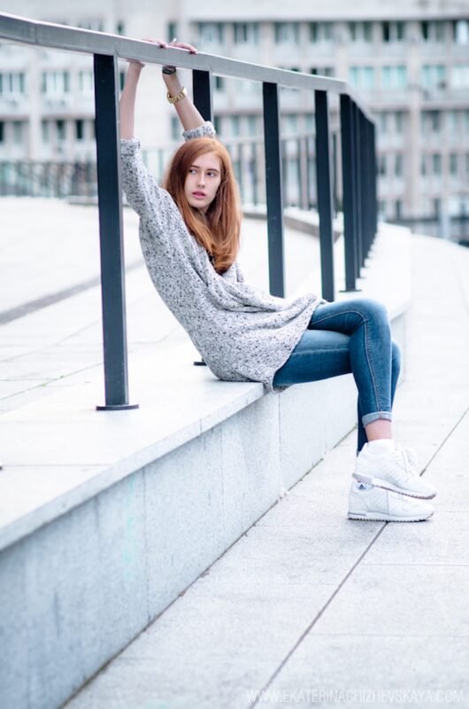 A young red-haired woman leaning back on a metal railing, wearing a grey knit cardigan, blue jeans, and white sneakers in an urban setting.