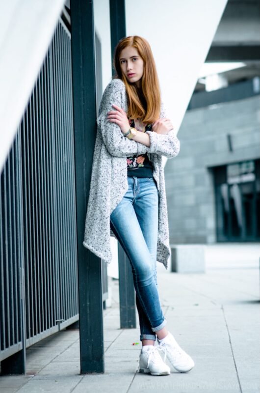 A full-length portrait of a red-haired woman with crossed arms, wearing a long grey cardigan, skinny jeans, and white sneakers, leaning against a pillar.