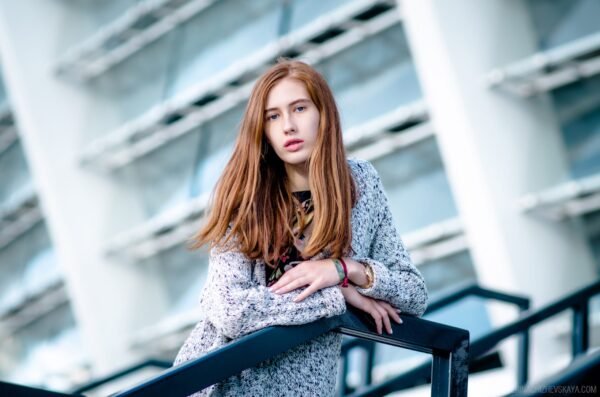 Close-up portrait of a red-haired woman leaning on a black handrail, wearing a speckled grey knit sweater with a modern building background.