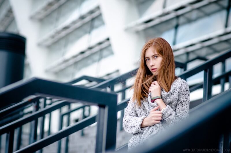 A young woman with straight red hair looking at the camera, pulling her grey cardigan close while standing on outdoor architectural stairs.