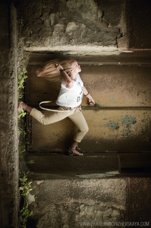 An expressive side view of a blonde woman with flowing hair, leaping horizontally inside a rustic concrete tunnel or alcove.