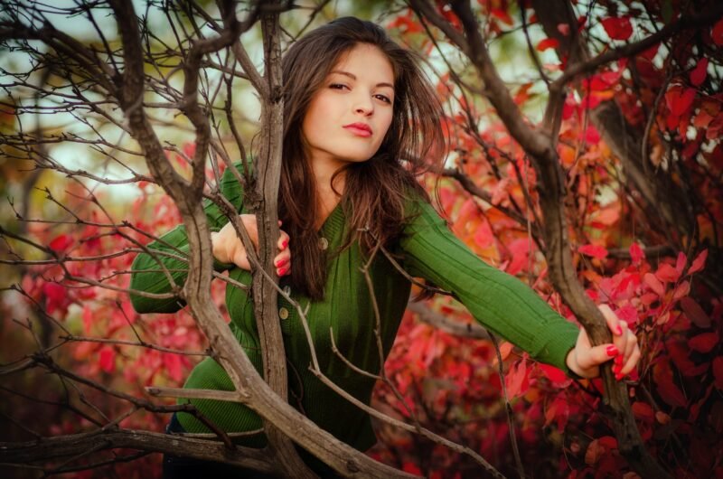 Beautiful woman in a green top looking confidently at the camera while standing behind autumn branches with red leaves.