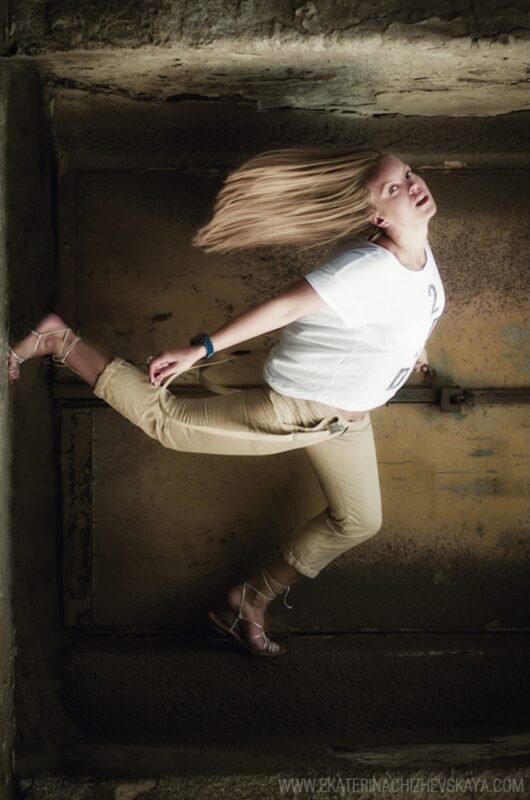 Dynamic action shot of a blonde woman with flying hair, wearing a white t-shirt and beige pants, posing in mid-air inside a concrete structure.
