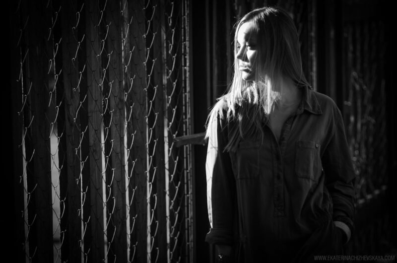 Dramatic black and white portrait of a blonde woman standing in low light next to a wire fence, looking to the side with deep contrast shadows on her face