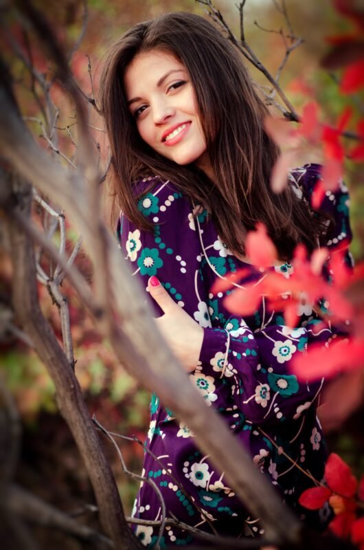 Smiling woman in a purple floral dress posing among tree branches and red autumn foliage.