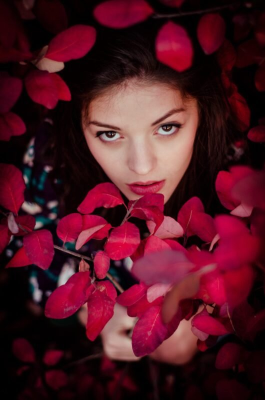 High angle close-up portrait of a woman looking up through vibrant red autumn leaves.