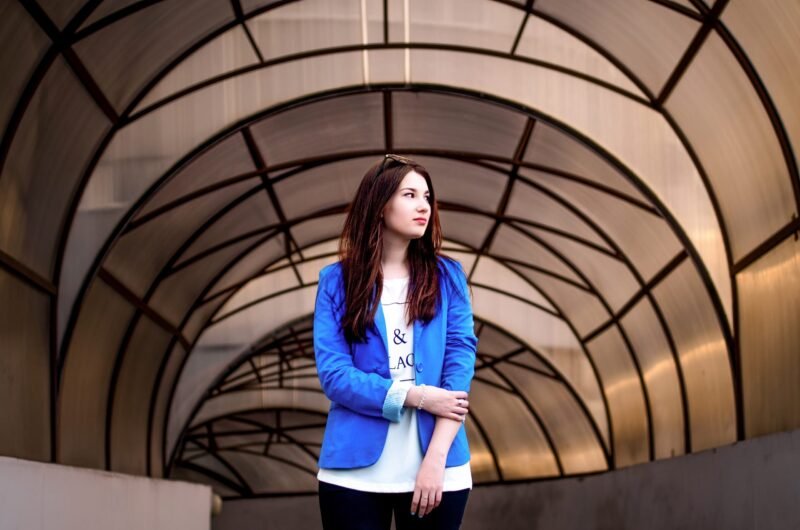 Young woman in a bright blue blazer standing and looking away under a long modern arched tunnel canopy.