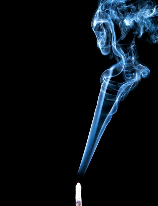 Thin stream of blue smoke rising straight up from a lit incense stick before expanding on black background